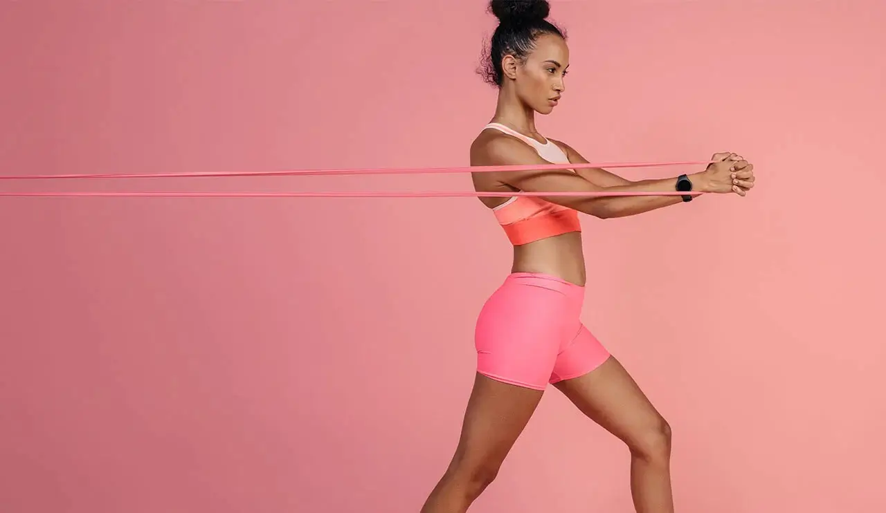 woman using exercise bands with pink shorts and top on pink background